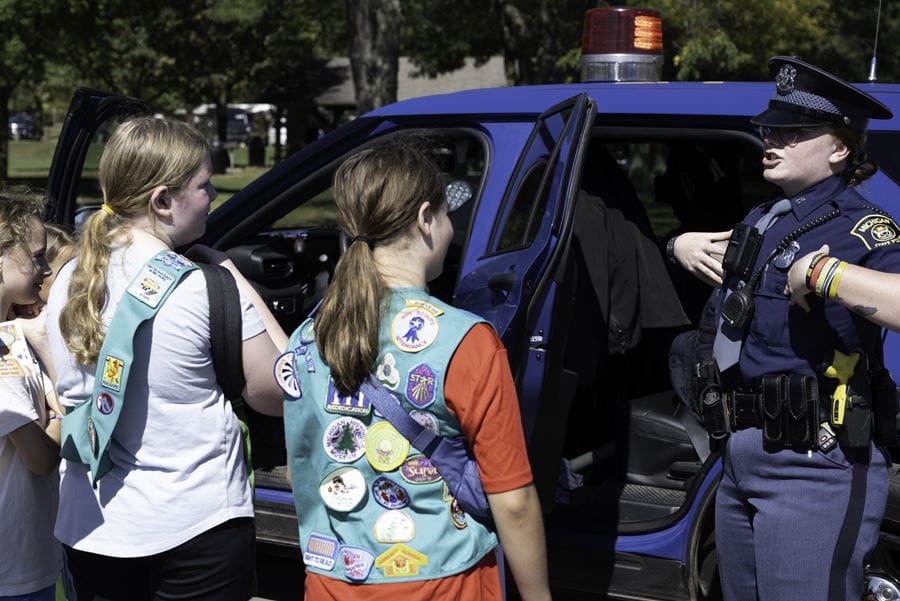 Two young girls wearing green girl scout vests and patches learning from an MSP trooper while standing next to a patrol vehicle.