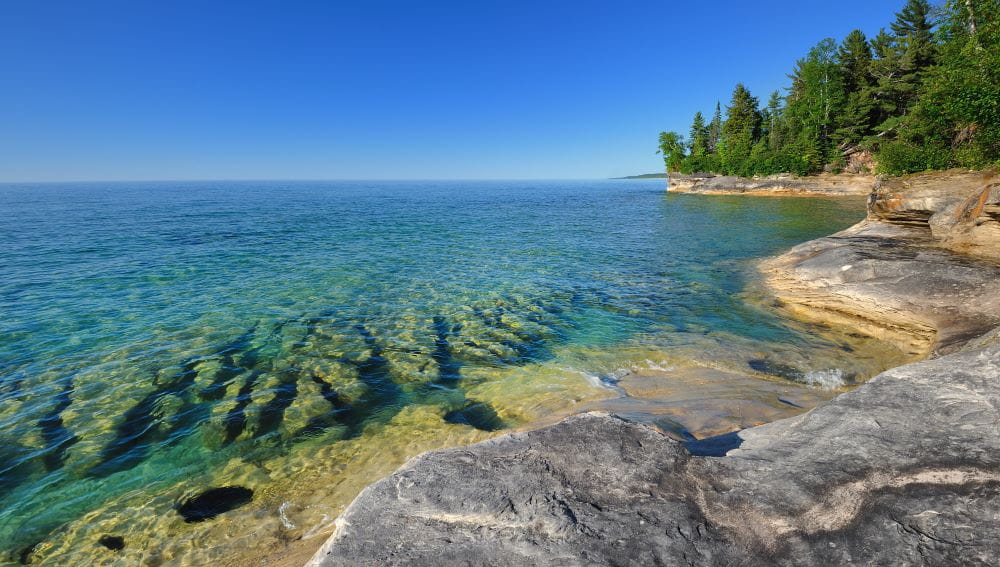 An endless expanse of turquoise-blue clear waters under a bright blue sky. The shoreline is made of large, smooth rocks.