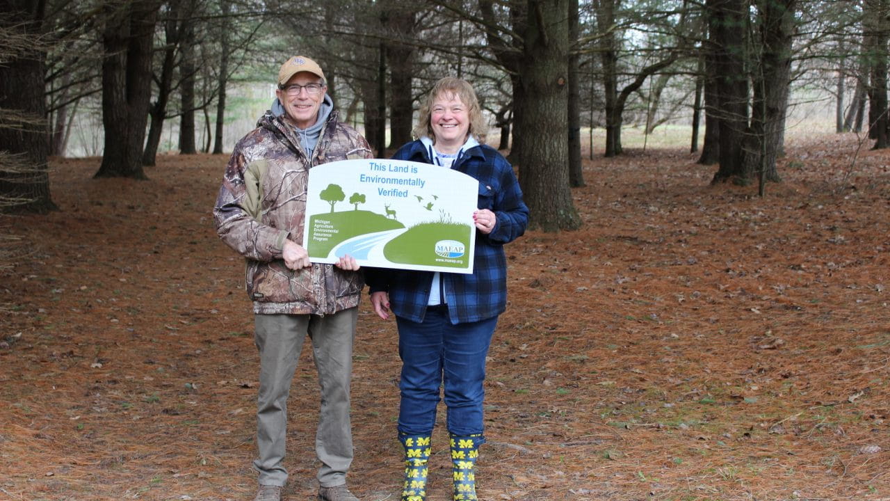 A couple in an evergreen forest holding a sign that says 'This Land is Environmentally Verified'