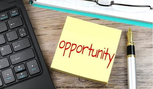"Opportunity" written in red ink on a yellow Post-It note on a desk next to a laptop and pen with paper