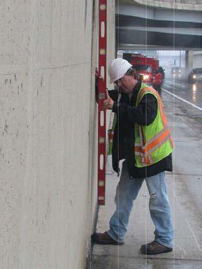 An MDOT worker holds a level against a retaining wall to measure tilt.