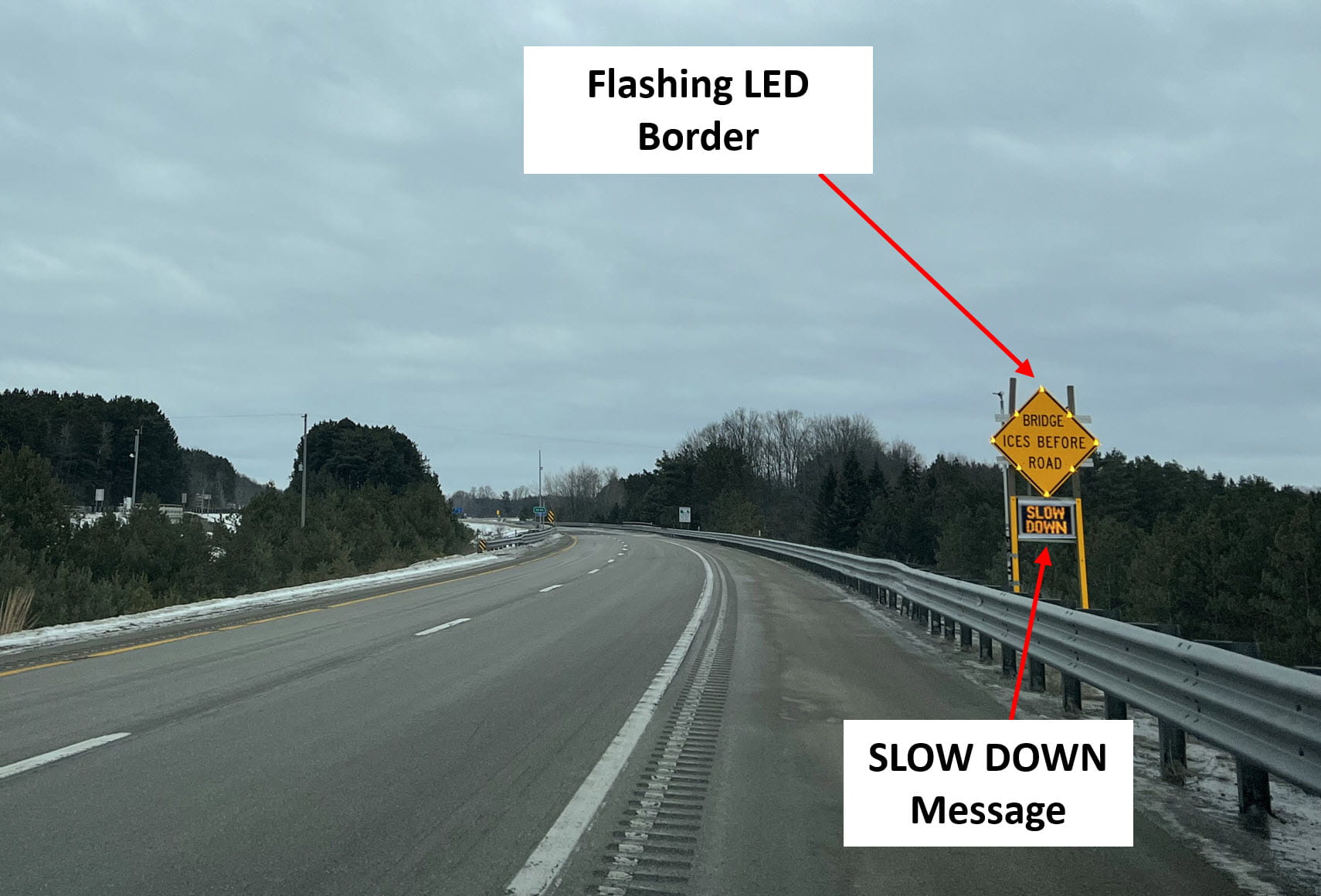 A weather-activated “BRIDGE ICES BEFORE ROAD” warning sign along a two-lane rural highway. The sign has a flashing LED border and a text panel below with the message “SLOW DOWN.”