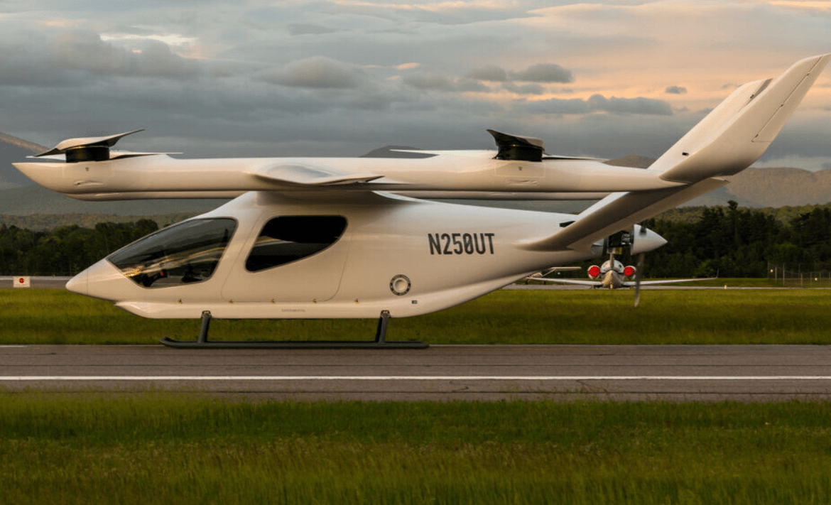 A white Beta Technologies ALIA aircraft on a runway with grass areas on either side and a cloudy sky.