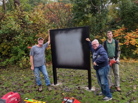 With trees in fall color, three men hold tools to install a sign frame. A generator and toolbox are in the foreground