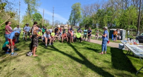 A woman in a blue shirt speaks to a crowd of 30 people on grass next to a trail