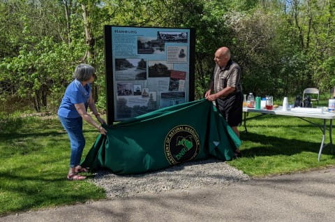 A woman and a man pull down a green drape with a Michigan DNR logo, revealing an interpretive sign in a black metal frame