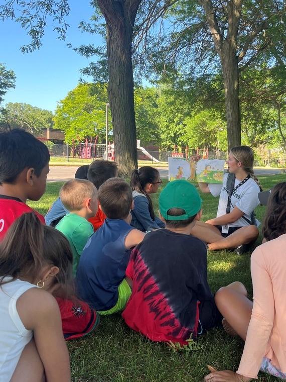 Children sitting on the grass in front of a counselor reading a book at an Outdoor Discovery Center event.