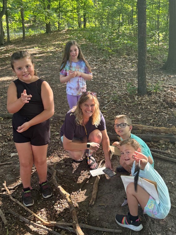 A teacher surrounded by four children in the woods smiling at the camera