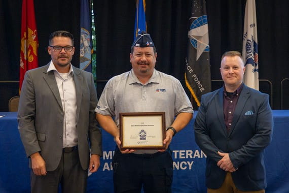 three light skinned men on stage with one in uniform holding a framed certificate